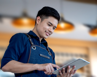 Seorang laki-laki sedang duduk dan menulis di meja sambil melihat ke arah tablet.