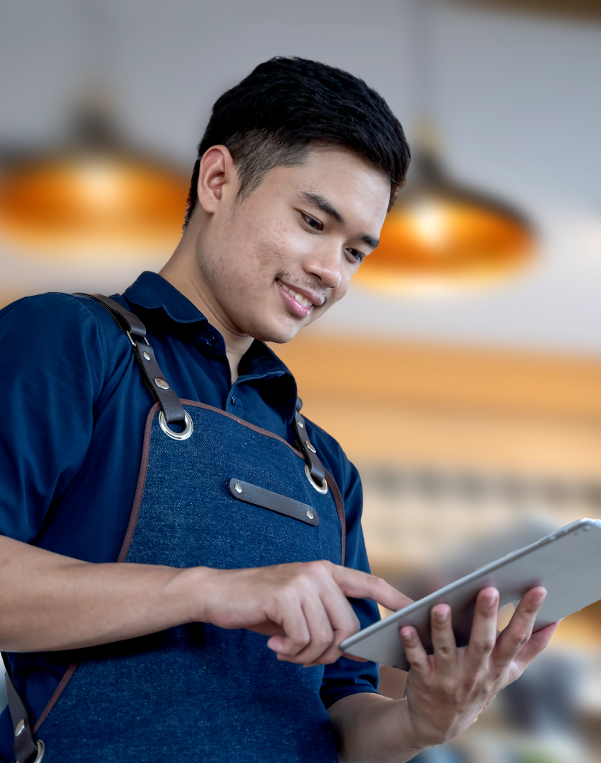 Seorang laki-laki sedang duduk dan menulis di meja sambil melihat ke arah tablet.